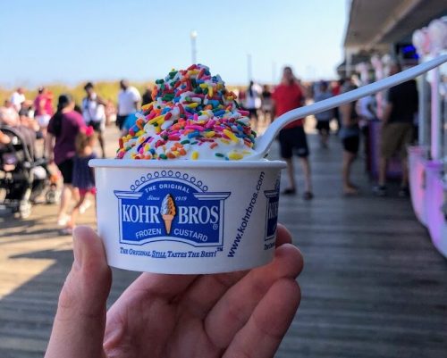A hand holds a cup of frozen custard topped with colorful sprinkles, with a busy boardwalk scene in the background.