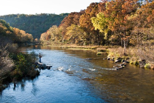 A serene river flows through a colorful autumn landscape, with a person fishing on a rocky bank.