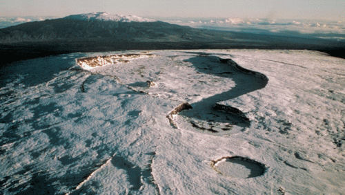 Aerial view of a snow-covered mountain landscape with glacial features and distant peaks under a clear sky.