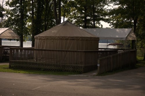 A tan yurt surrounded by wooden decking, near a lake, with trees in the background and a white tent nearby.