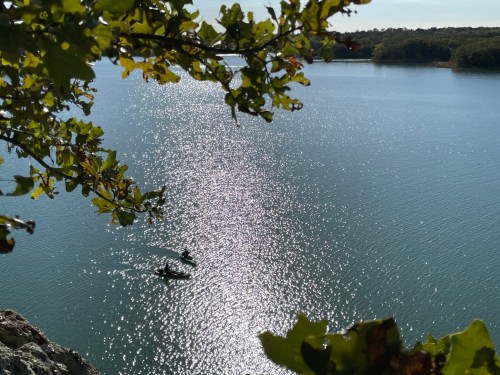 Two kayakers paddle on a shimmering lake, framed by green leaves and a clear blue sky.
