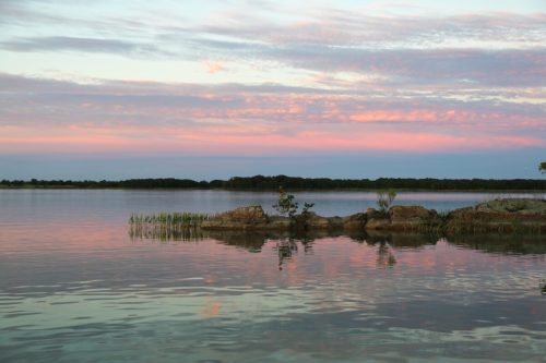 A serene lake at sunset, with soft pink and purple clouds reflecting on the water and rocky islands in the foreground.