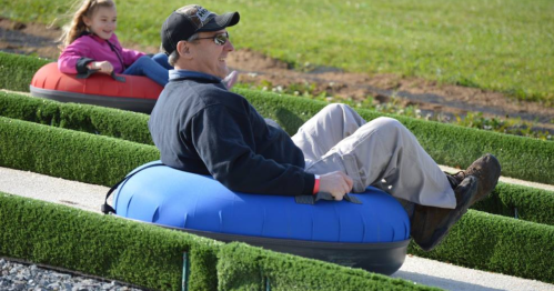 A man and a girl enjoy tubing down a grassy slope on inflatable tubes, smiling and having fun outdoors.
