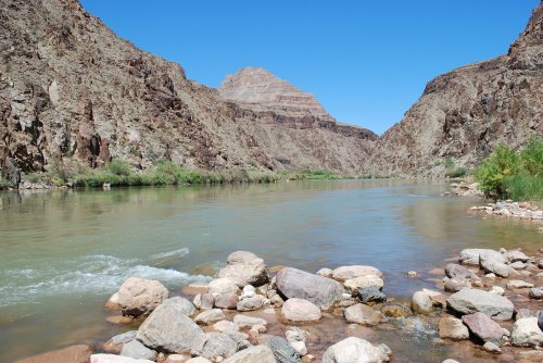 A serene river flows through rocky canyon walls under a clear blue sky, with greenery along the banks.
