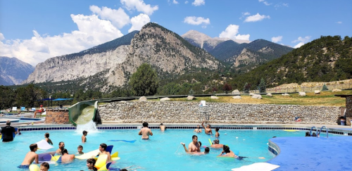 A lively pool scene with children playing, surrounded by mountains and a clear blue sky.