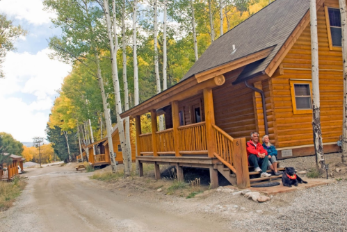 Two people sit on the porch of a log cabin, with a dog resting nearby, surrounded by trees and a dirt road.