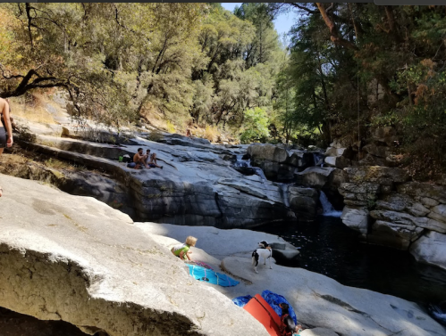 A rocky riverbank with people relaxing by the water, surrounded by trees and a clear blue sky.