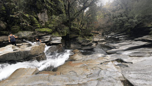 Two people explore rocky terrain beside a flowing river, surrounded by lush greenery and trees.