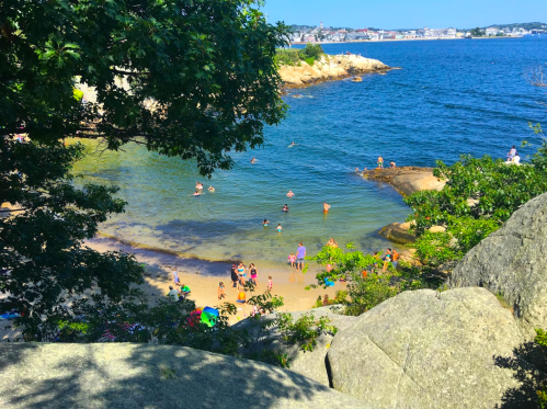 A scenic view of a beach with people swimming and relaxing, surrounded by rocks and greenery under a clear blue sky.