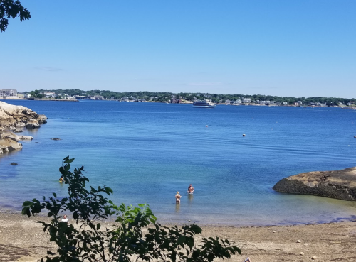 A serene beach scene with clear blue water, a few people wading, and distant boats under a bright blue sky.