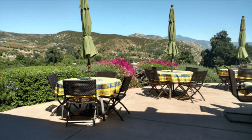 Outdoor patio with tables and chairs, surrounded by greenery and mountains, under clear blue skies.