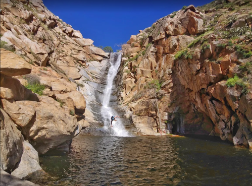 A waterfall cascades down rocky cliffs into a serene pool surrounded by rugged terrain and greenery.