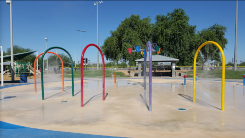 Colorful water play structures in a sunny park, with mist spraying and a picnic area in the background.