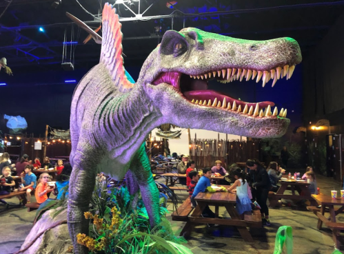 A large dinosaur statue with vibrant colors looms over children dining at picnic tables in a themed restaurant.