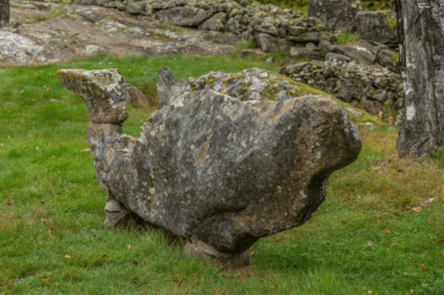 A large, weathered stone sculpture resembling a whale, set in a grassy area with rocky terrain in the background.