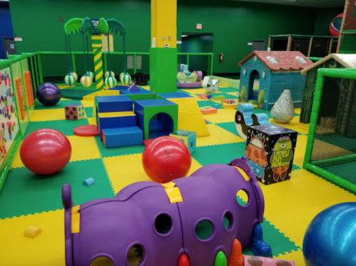 Colorful indoor play area with soft structures, balls, and playhouses for children. Brightly colored flooring and walls.