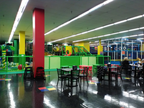 Brightly colored indoor play area with climbing structures, tables, and chairs. Large windows let in natural light.