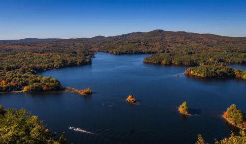 A scenic view of a tranquil lake surrounded by autumn-colored trees and rolling hills under a clear blue sky.