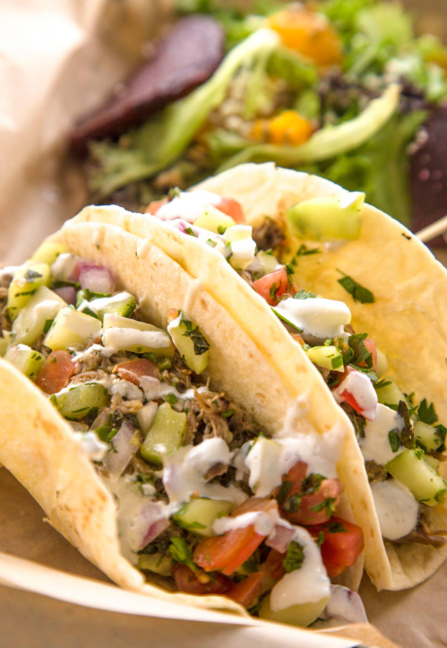 Three tacos filled with diced vegetables, herbs, and sauce, served with a side salad on a brown paper tray.