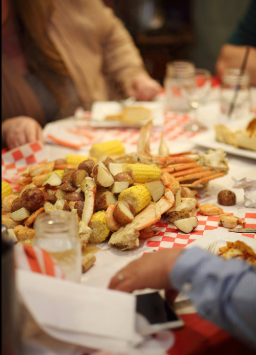 A table spread with a seafood feast featuring crabs, corn, potatoes, and sausage, surrounded by diners.