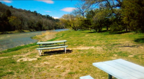 A serene riverside scene with picnic tables on grassy land, surrounded by trees and a clear blue sky.