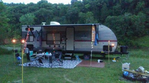 A parked camper trailer in a grassy area, surrounded by trees, with outdoor seating and string lights.