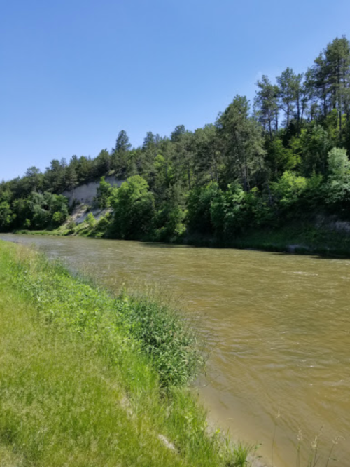 A river flows through a lush green landscape, bordered by trees and rocky cliffs under a clear blue sky.