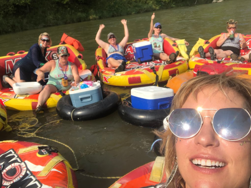 A group of six women enjoying a fun day floating on colorful inner tubes in a river, smiling and waving at the camera.