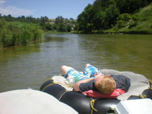 A person relaxes on a floating tube in a calm river, surrounded by greenery and blue skies.