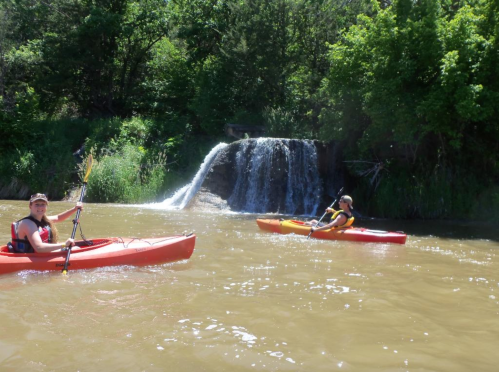 Two people kayaking on a river near a small waterfall, surrounded by lush greenery on a sunny day.
