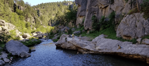 A serene river flows through rocky terrain, surrounded by lush greenery and people enjoying the natural landscape.