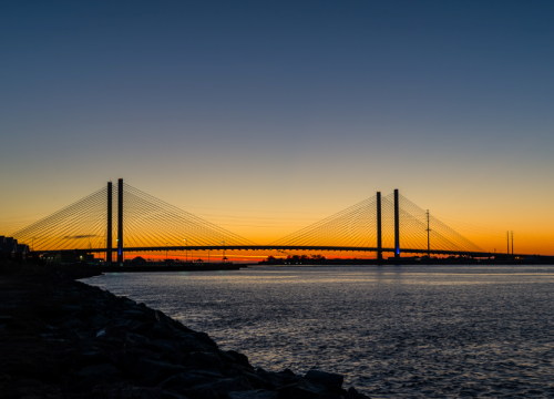 Silhouette of a cable-stayed bridge at sunset, with a calm river and colorful sky in the background.