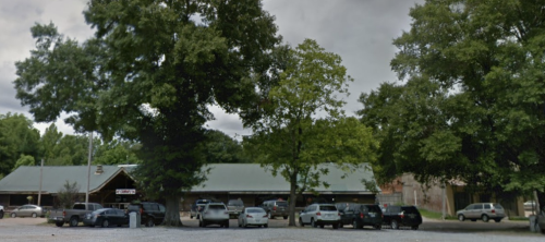 A parking lot with several cars, surrounded by trees and a building with a green roof in a cloudy sky.