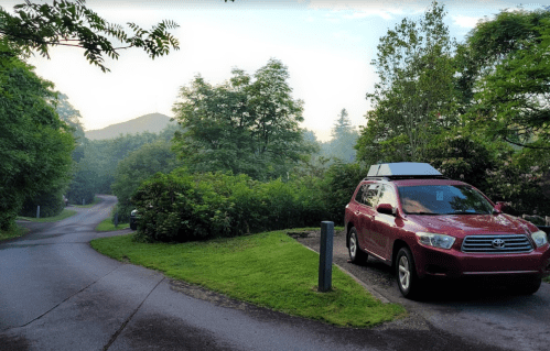 A red SUV parked on a winding road surrounded by lush greenery and trees, with a misty mountain in the background.