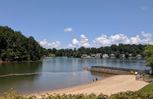 A serene lake scene with a sandy beach, wooden dock, and houses in the background under a blue sky with fluffy clouds.