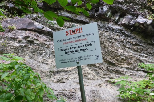 Sign warning against climbing rocks near a waterfall, stating it's dangerous and has led to fatalities.