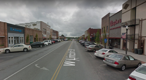 A street view of W. Lincoln St. lined with parked cars and storefronts under a cloudy sky.