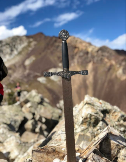 A sword stands upright on rocky terrain, with mountains and blue sky in the background.