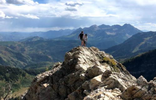 Two hikers stand on a rocky peak, overlooking a vast mountain landscape under a cloudy sky.