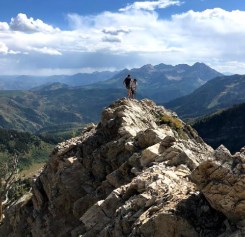 A person stands on a rocky mountain peak, overlooking a vast landscape of mountains and valleys under a blue sky.