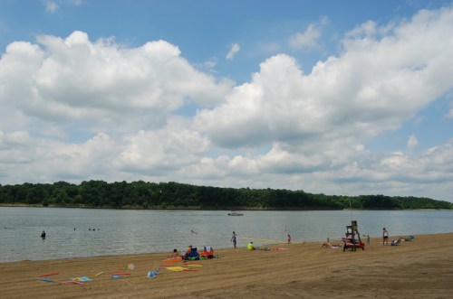A sandy beach by a calm lake, with people enjoying the water and clouds in the sky. Green trees line the shore.