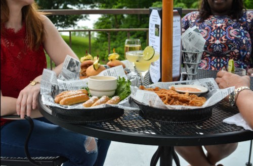 A table with two baskets of food, including hot dogs and fried items, alongside drinks and a menu in a casual outdoor setting.