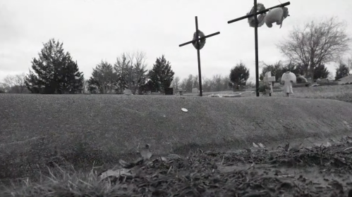 A black and white image of a cemetery with two crosses and a small figure in the background among bare trees.