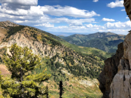 A scenic view of mountains and valleys under a blue sky with scattered clouds. Lush greenery covers the slopes.