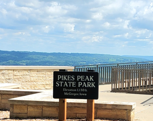 Sign for Pikes Peak State Park in McGregor, Iowa, with a scenic view of hills and a partly cloudy sky.