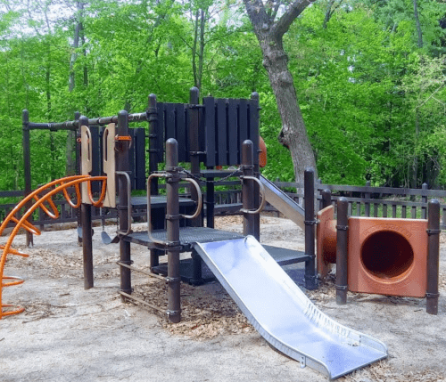 A playground structure with a slide, climbing bars, and a tunnel, surrounded by trees and gravel ground.