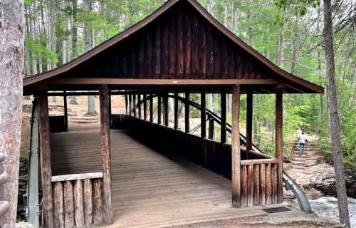 A wooden covered bridge surrounded by trees, with a person walking near a stream in the background.