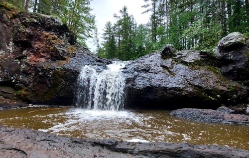 A small waterfall cascades over dark rocks into a serene pool, surrounded by lush green trees.