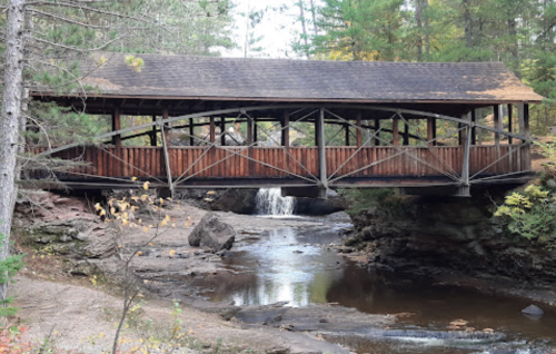 A wooden covered bridge spans a creek, surrounded by trees and rocky terrain, with a small waterfall nearby.