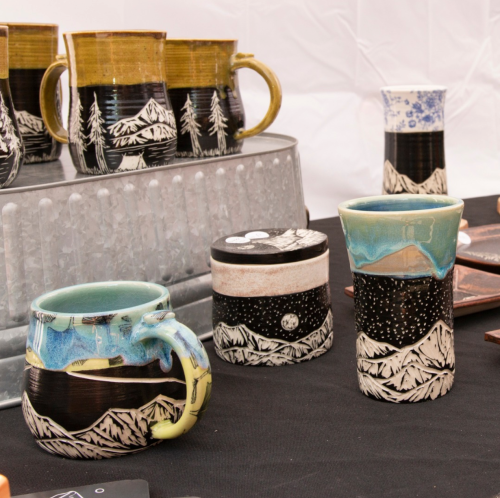 A display of handcrafted ceramic mugs and containers with mountain and nature designs on a black tablecloth.